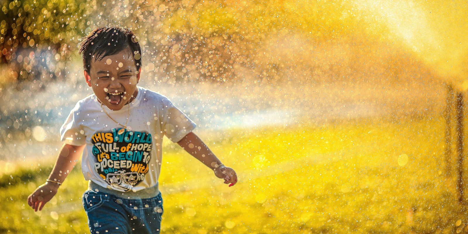A young Asian boy with short hair, a graphic t-shirt and jeans smiles as he runs through a sprinkler with golden light and water splaying behind him.