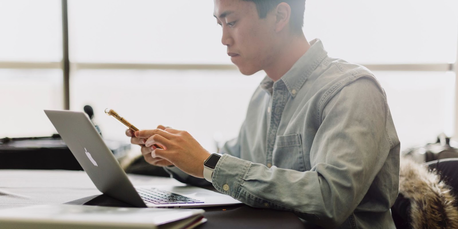 A young Asian man in a button down shirt sits at his desk, looking into his phone, with a laptop open in front of him.