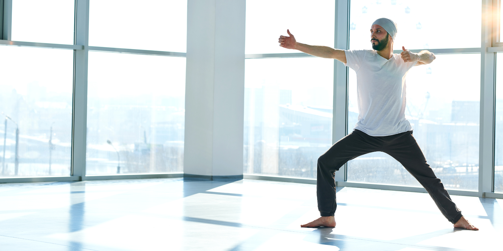 A man with a short beard wearing black workout pants, a white t-shirt and a white turban does a yoga warrior pose in an empty room with large windows overlooking a cityscape.