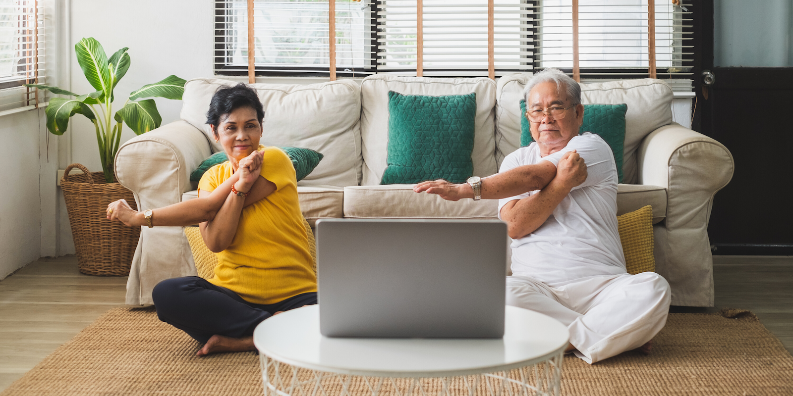 A senior Asian woman with short black hair wearing black pants and an orange t-shirt and senior Asian man with white hair wearing white pants and a white t-shirt sit on the floor in front of a couch, an open laptop on the table in front of them. The sit cross-legged and stretch their arms across their chests following the program on the computer.
