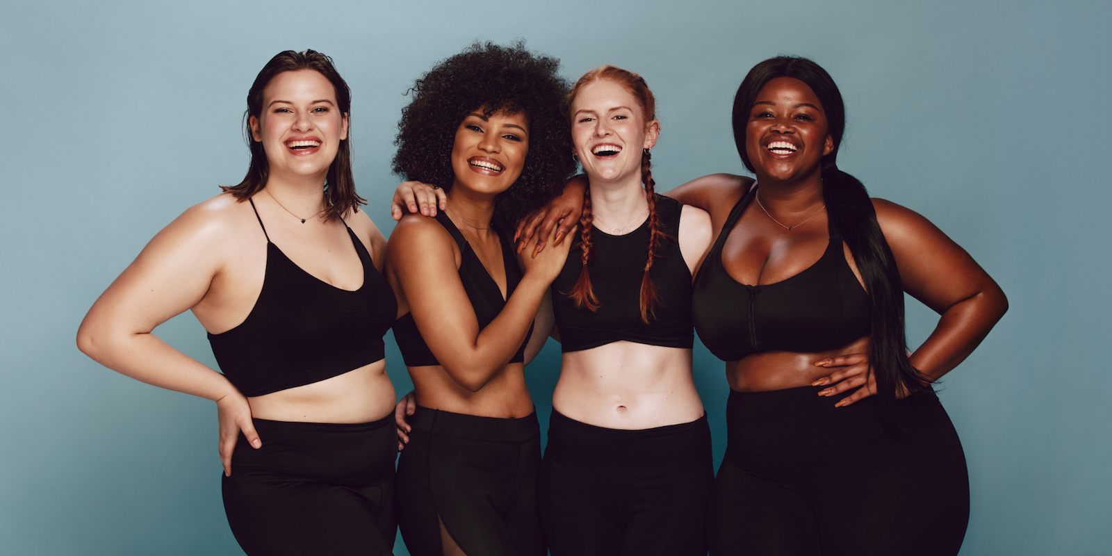 Four young women in black fitted yoga clothes stand against a blank blue wall linking arms and smiling into the camera: A white woman with medium-length brown hair, a Black woman with a brown Afro, a white woman with long red braids, and a Black women with her long dark hair pulled behind her and draping down her side.
