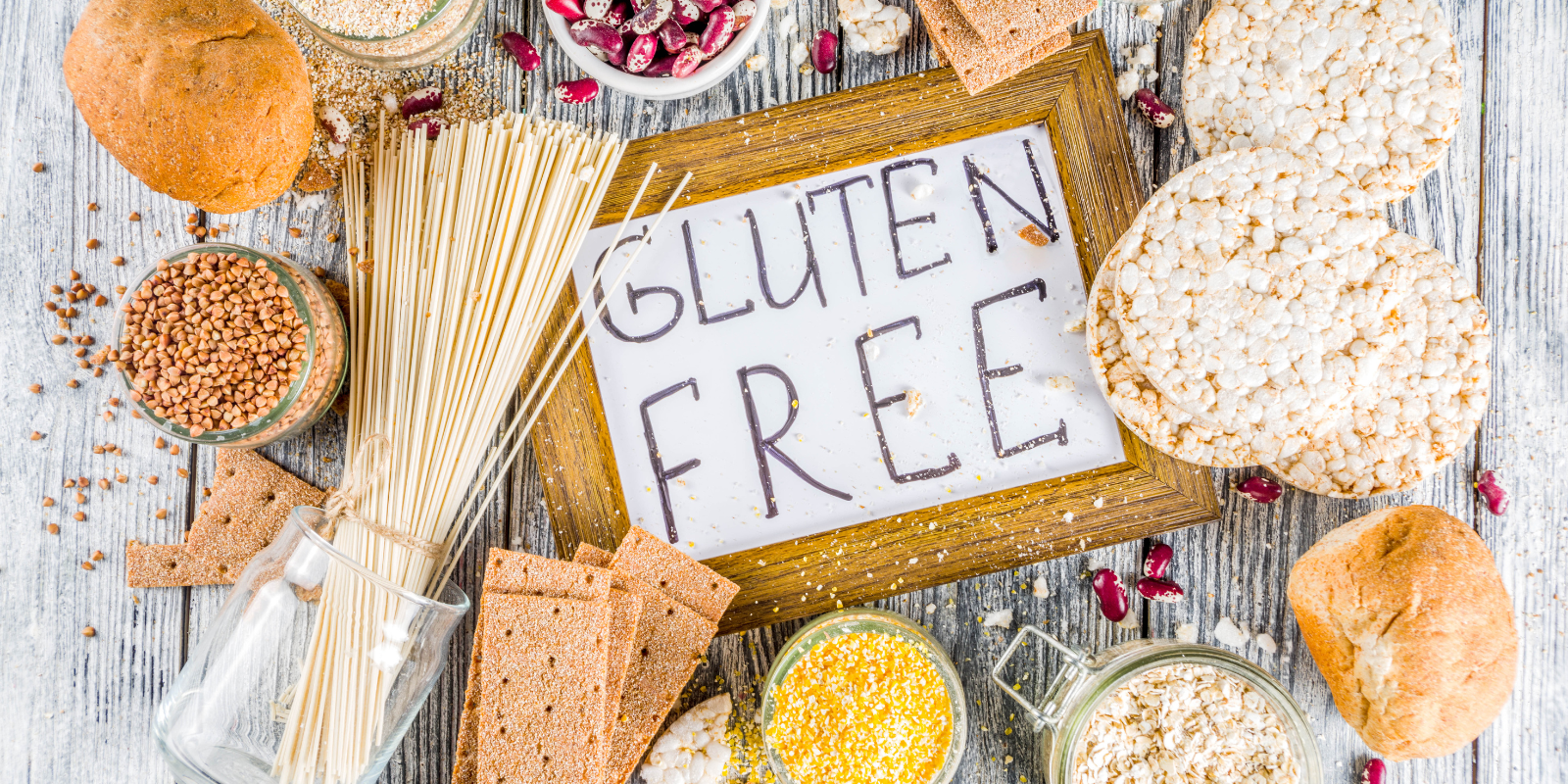 A collection of gluten-free items on a gray wood background, things like rice cakes, bowls of seeds, etc. A whiteboard sign in the middle says "gluten FREE".