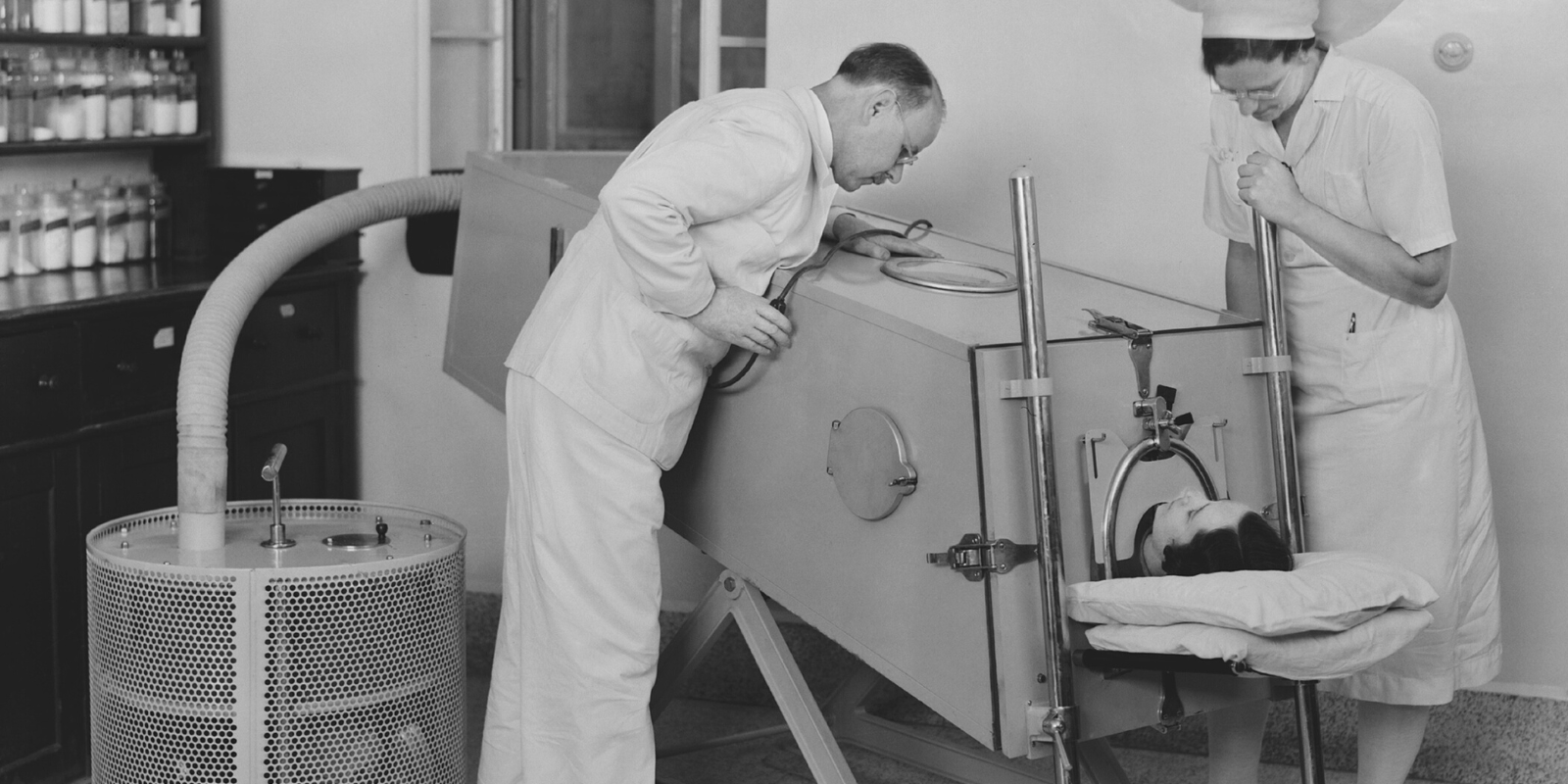 A black and white photograph from around the 1950s, with a doctor and nurse looking over a polio patient in an iron lung machine.