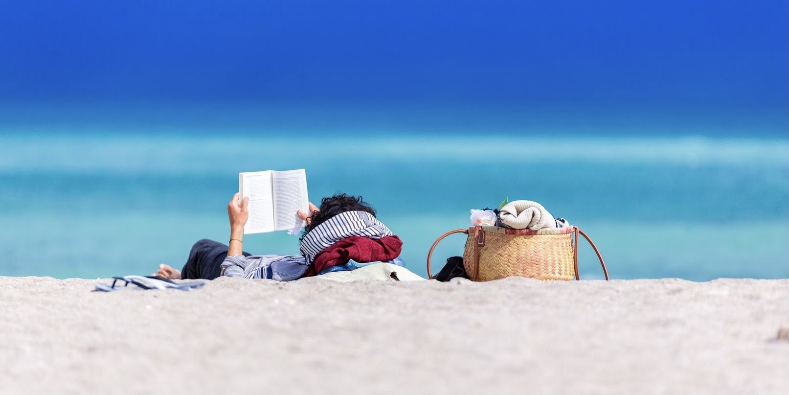 A photograph of a white woman laying on beach sand with a basket/purse nearby, reading a book.