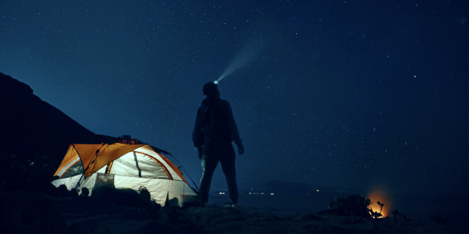 A photograph of a night scene with a tent lit up from inside, a campfire burning down, and a man standing with a headlamp lighting a beam into the dark sky.