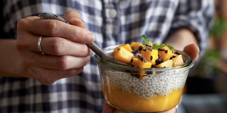 A close up photograph of the torso and arms of white person in a checkered shirt holding a glass dish of chia pudding and mango.