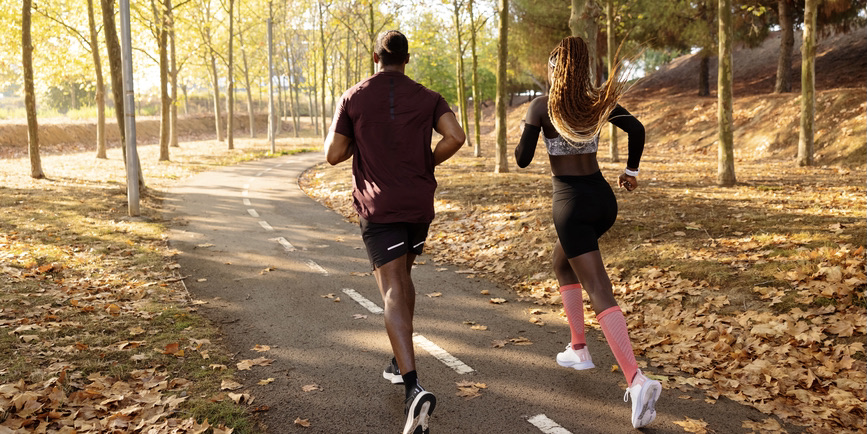 A color photograph of a Black man and Black woman taken from behind They're in workout clothes jogging along a path in a park flanked by fallen yellow leaves.