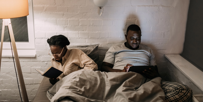 A color photograph of a Black man and Black woman in a bed against a white painted brick wall with gray bedsheets around them. The woman is reading a book and the man gazing into a lit screen in his lap.