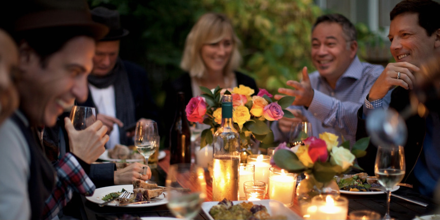 A cozy outdoor color photograph of a group of 40-something friends eating at a table with low candles and flowers.