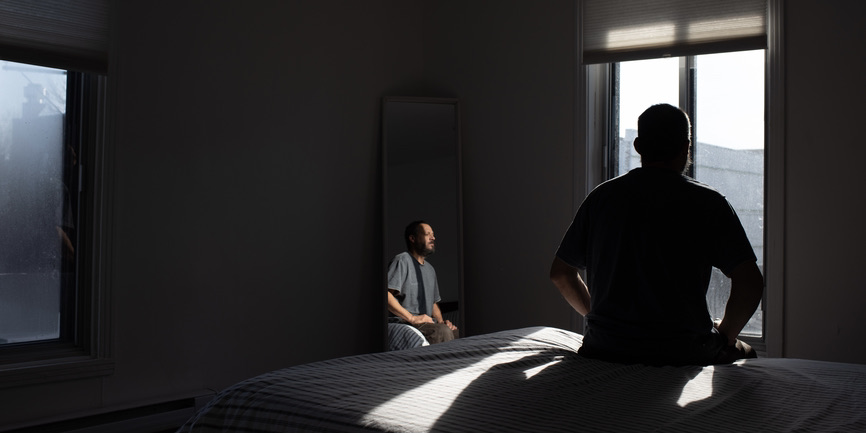 A color photograph of a man sitting on a bed with a gray coverlet, his back to the camera, looking out a window. Much of the room is in shadow. And a mirror in the corner reflects his image back.