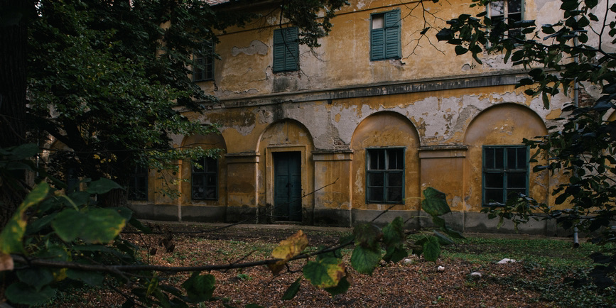 A color photograph of the exterior of an abandoned hospital, with orange stucco walls and vines growing up the side.