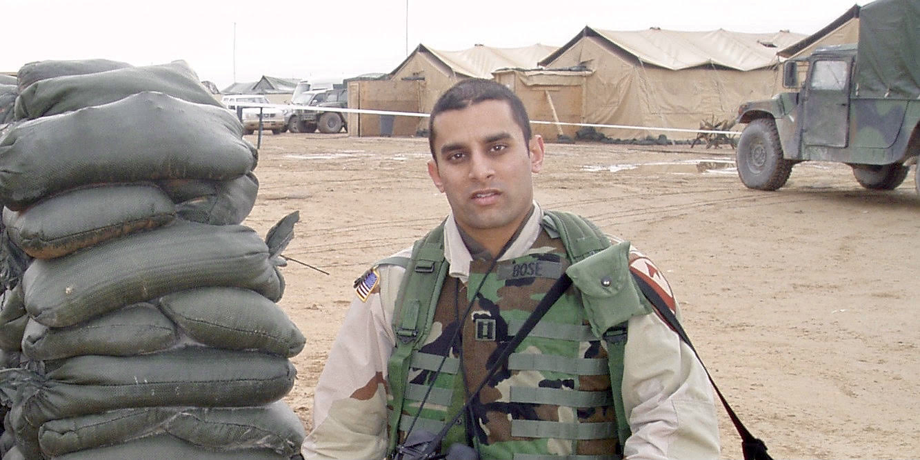 A color photograph of Dr. Sudip Bose, an Indian American combat physician with short dark hair in military fatigues. He's standing in a desert in front of a line of tan tents and a military vehicle.