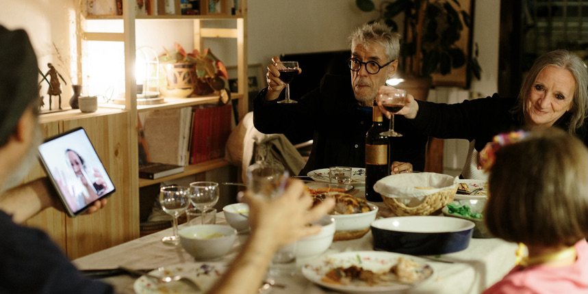 A color photograph of a multi-generational white family gathered at the dining room table, cheersing to someone on a video screen.
