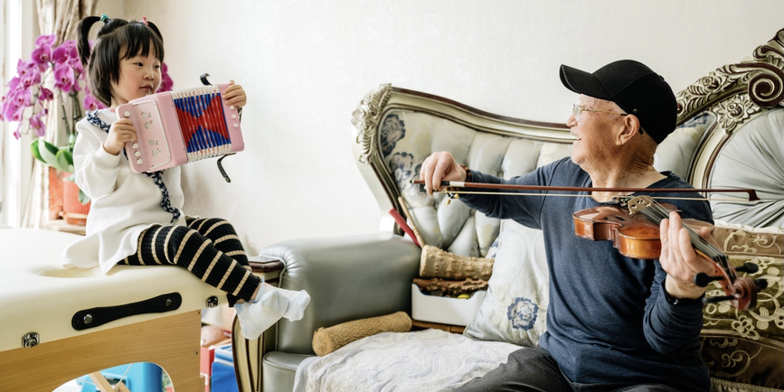 A color photograph of an Asian senior man wearing a blue shirt and a black baseball cap, playing a violin and smiling at his granddaughter, a young Asian girl with short black hair wearing a white and black outfit and playing a small accordion. 