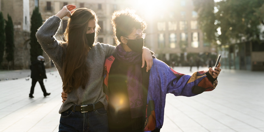 A color outdoor photograph of a young white woman with long brown hair and a young Black woman with her black hair tied up. They're wearing long sweaters and black N95 masks, cuddling together as they take a selfie.