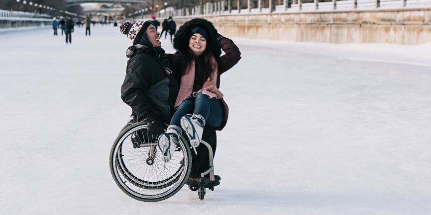 An outdoor color photograph of a frozen river on which a white man in a wheelchair wearing a red pompom hat laughs with a white woman wearing a dark hooded coat and pink scarf sits on his lap and laughs, too.