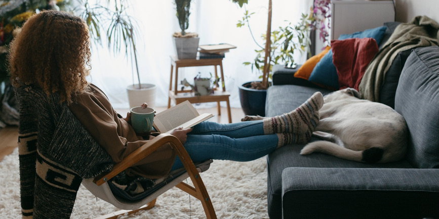 A color indoor photograph of a young Black woman wearing jeans and a sweater, her full brown hair hiding her face, reading a book with her legs and socks perched on a blue couch where a dog lays sleeping.