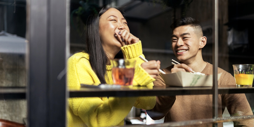 A color photograph taken through a window into a restaurant, where a young Asian couple wearing long sweaters sit eating food from a paper tray with chopsticks, smiling and laughing.