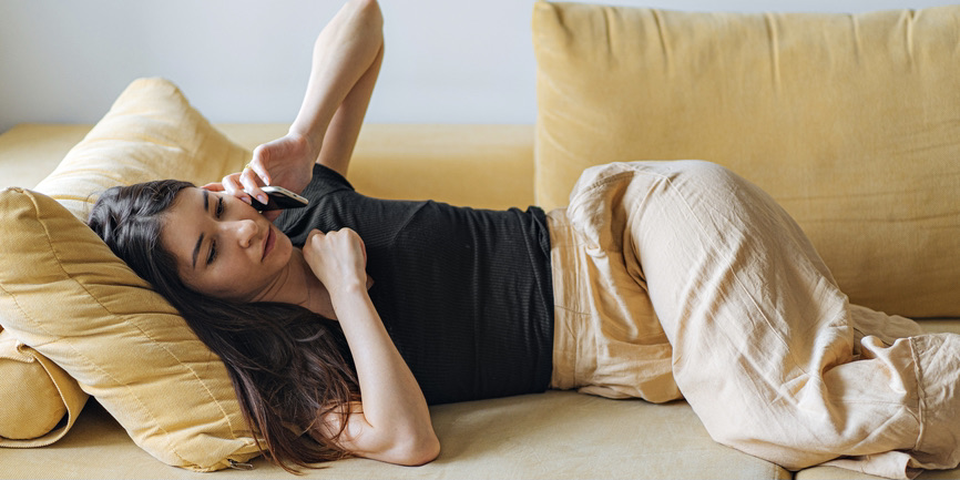 A color indoor photograph of a young woman with long black hear, wearing a black t-shirt and linen pants, laying on a gold-colored couch, talking into her phone.