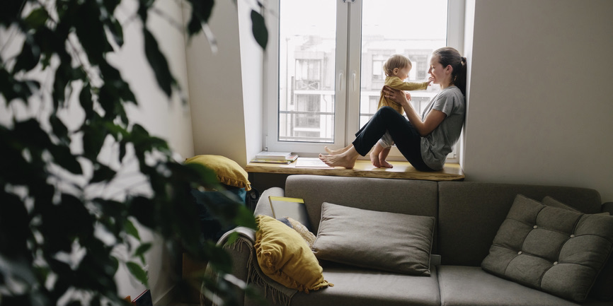A color indoor photograph of a white woman wearing a gray shirt and jeans, sitting in a windowsill playing with her toddler. Closer to the camera is a gray couch with gold pillows and a large palm plant.