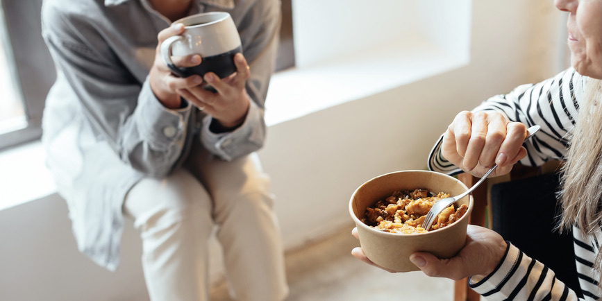 A closeup color photograph of two women, perched on a chair and a windowsill, one drinking out of a mug and the other having cereal from a bowl.