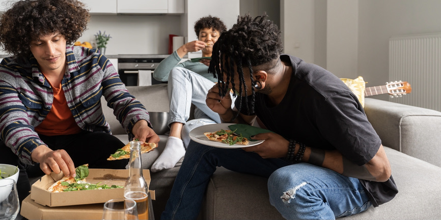 A color indoor photograph of three Black teens sitting on couches around a coffee table, digging into a pizza topped with green vegetables.