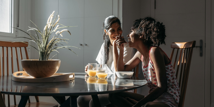 A color indoor photograph of a mother and daughter with dark skin sitting at a breakfast table in sunlight, eating breakfast, talking and smiling.