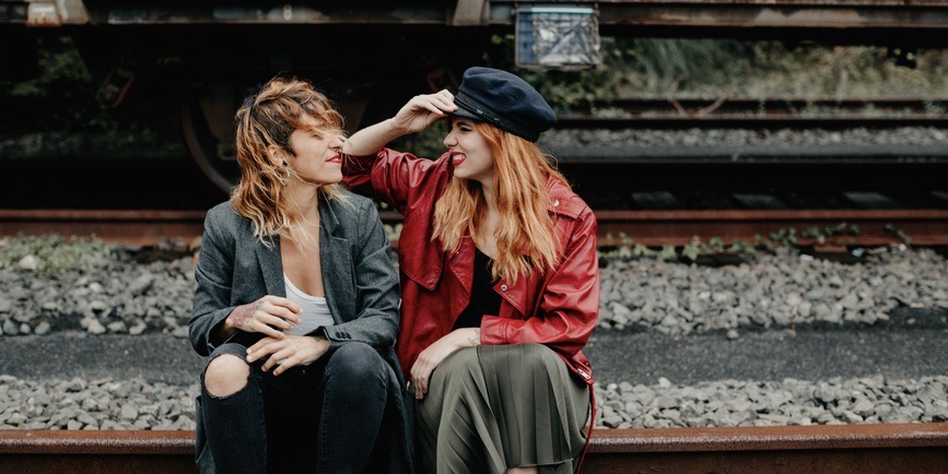 A young lesbian couple sit outdoors on a set of train tracks. Both are white with long red hair. One wears ripped jeans, a white tank and a gray blazer. The other a green skirt, red leather jacket and black cap. They're squinting and smiling at each other. 