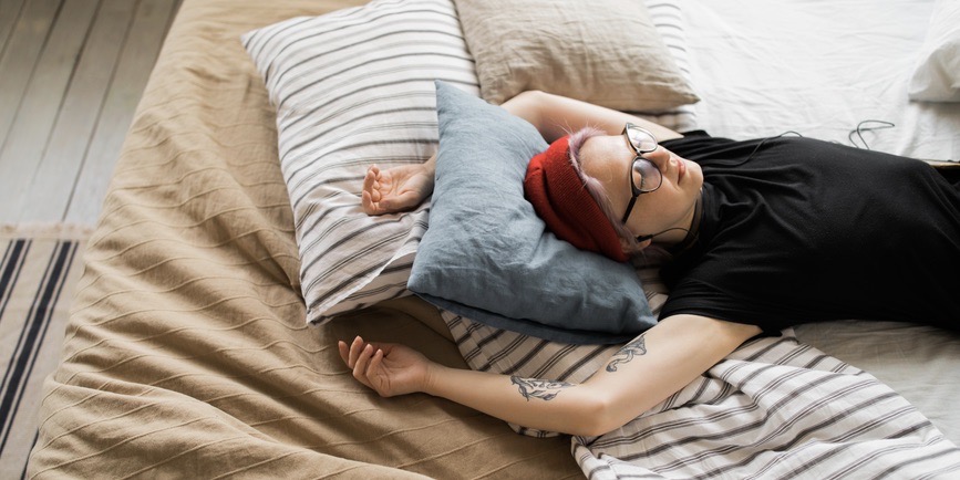 A color indoor photograph of a young white person with short pink hair hidden under a red cap wearing a black t-shirt and black-rimmed glasses lies on a bed covered with sheets and pillows. They're listening to music with their eyes closed, their tattooed arms stretched above them.