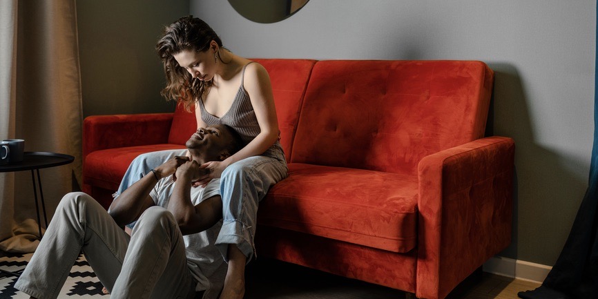 A dreamy color indoor photograph of a white woman with long brown hair wearing a tank and jeans sits on a bright red velvet couch, looking down into the face of her boyfriend, a Black man with short black hair wearing khaki pants and a white t-shirt, who holds her hands and gazes up at her. 