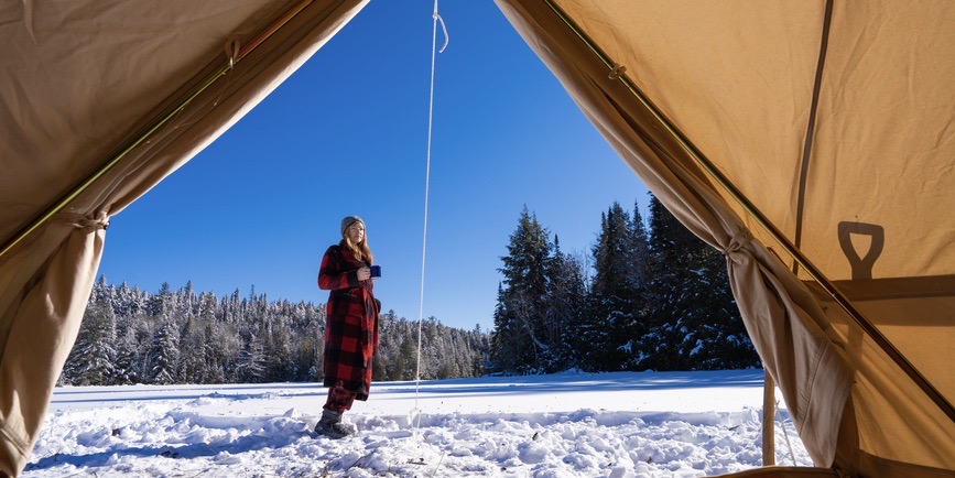 A color outdoor photograph taken from the inside of a tent looking out, where a young white woman wearing a red plaid robe stands in the snow against a blue sky and fir trees, drinking a cup of coffee.