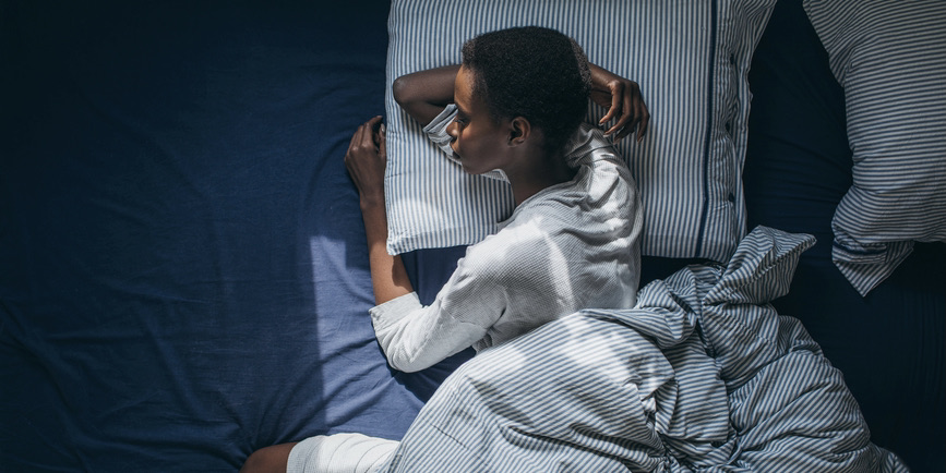 A thin Black woman with short black hair wearing white pajamas lays sleeping on a striped blue and white pillow and blue sheets.
