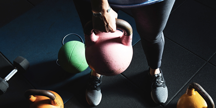 An overhead indoor color photograph of a white woman wearing gym clothes, holding a pink kettlebell weight, with more colorful weights on the floor below her.