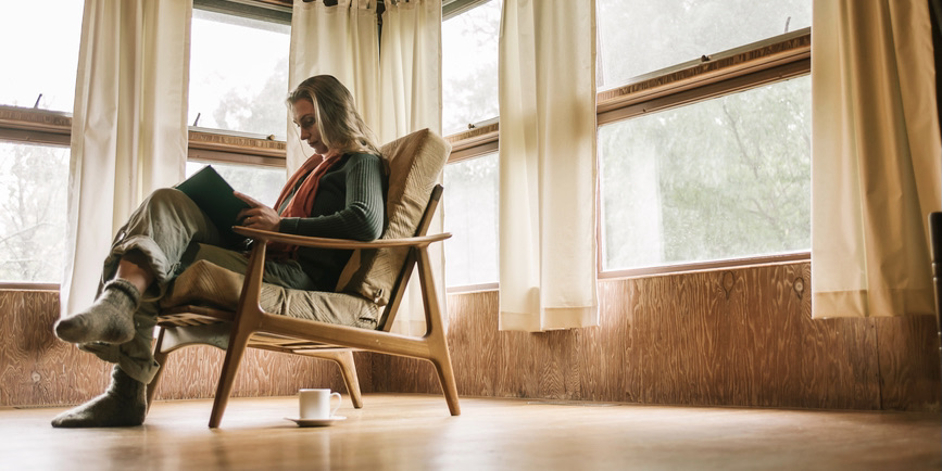 A color indoor photograph of a white woman with long blond hair wearing pants, a sweater, and thick socks sitting in an armchair reading a book in a sunny windowed room.