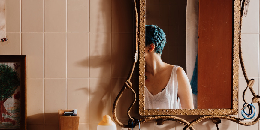 A color  The faceless reflection of a young white woman with blue hair seen in a bathroom mirror, with punk-hued wood plank walls of empty emerald green theater seats.