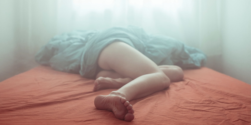 A blurry color photograph of a white person’s legs sticking out from below a light blue sheet, as they lay atop a pinkish-sheeted bed
