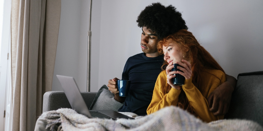 A young couple sits on a couch under a blanket, watching something on a laptop while holding mugs in their hands. The man is Black with black hair, wearing a black shirt. The woman is white with red hair, wearing a long-sleeved orange shirt.