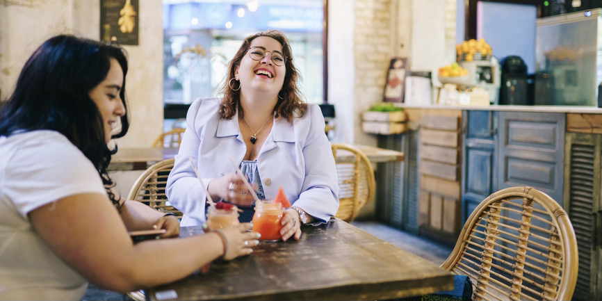 Two curvy white-skinned women sit at a table in a juice bar, holding glasses of juice in their hands. One woman with black hair looks down at the table as the other  with brown hair and glasses smiles with her face tilted upward.