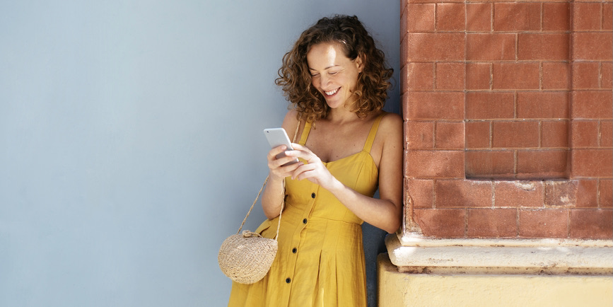 A young, thin white woman with curly brown hair wearing a yellow sundress stands against a blue wall and brick pillar, smiling as she looks down into her phone.