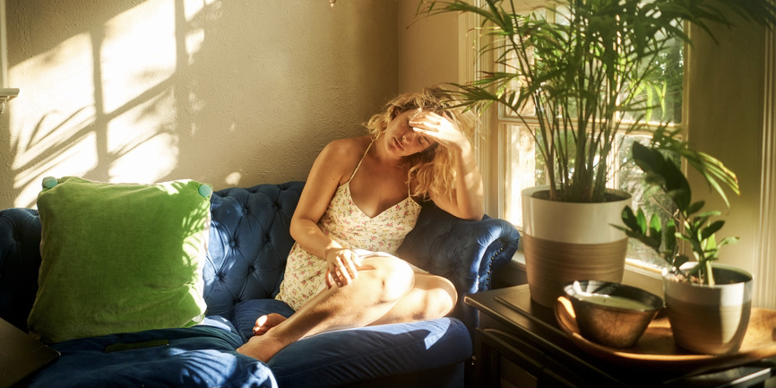 A color indoor photograph of a white woman with blond hair wearing a sundress. She sits on a blue couch with plants next to her, hiding her face from sunshine coming in through a window.