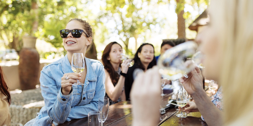 A colorful outdoor photograph of a wine tasting bar, with a young white woman sitting at front with her brown hair pulled up, wearing a jean jacket and sunglasses, smiling as she holds a glad of wine and looks off camera.