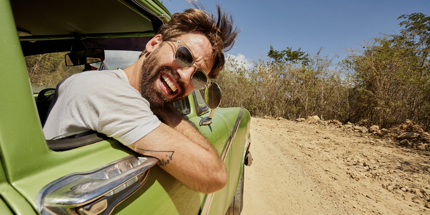 A color outdoor photograph of a young white man with brown hair and a beard wearing a white t-shirt, leaning out of a green vintage car window and smiling into the camera.