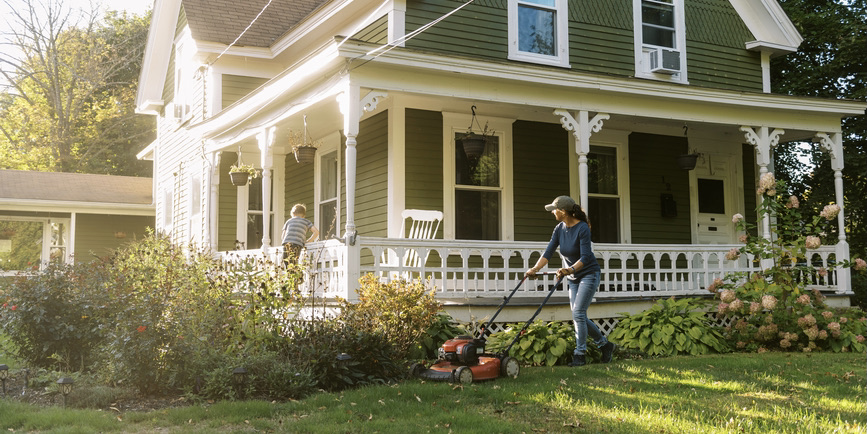 A color outdoor photograph of a middle-aged white woman weaing jeans and a blue shirt, moving the lawn outside her colonial house, while her son climbs around the porch.