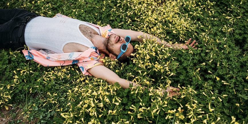 A colorful outdoor photograph of a young white man with a short beard, blue sunglasses and a colorful pink shirt, lying on a patch of yellow flowers, arms up and facing the sky.