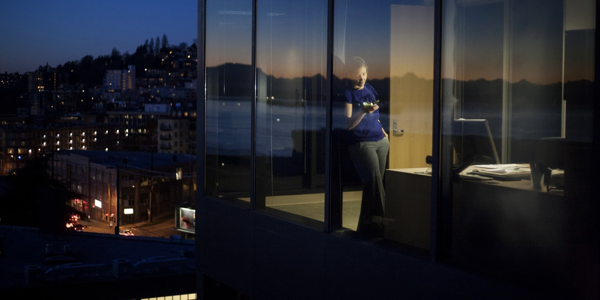 View looking into a window of a white female executive standing next to large office windows using a cell phone at sunset in Seattle, Washington, USA