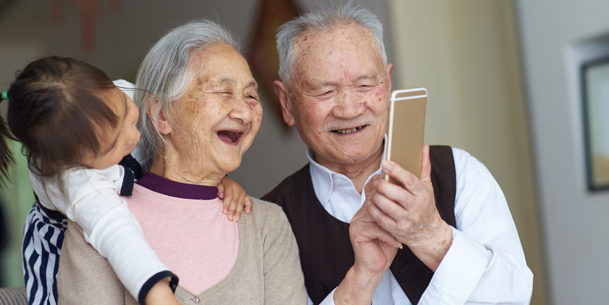 A happy senior Asian couple indoors with a small child on their shoulder laugh into a smart phone.
