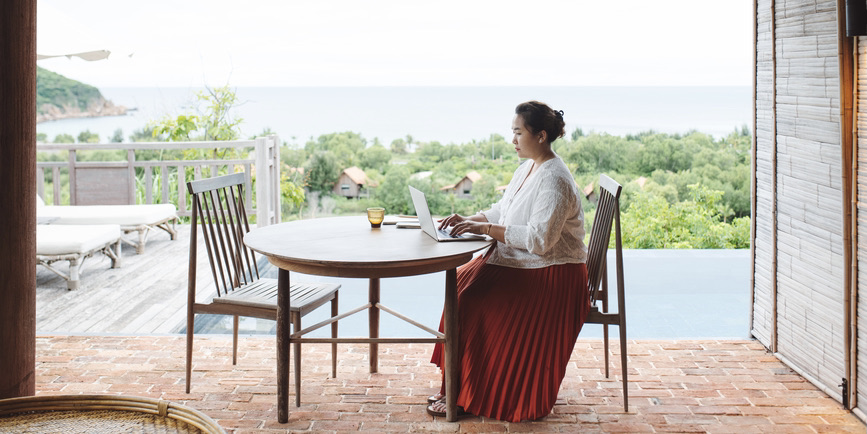 A beautiful, curvy Asian woman wearing a red skirt and white blouse types on laptop in the morning, sitting outside on a deck with a view of water in the distance. 