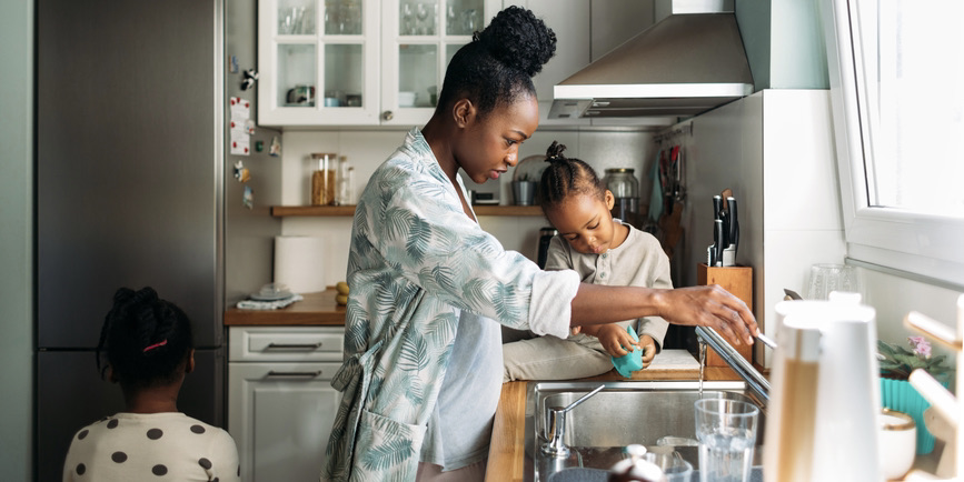 A Black woman wearing a white and blue robe and her little daughters wash dishes in a sunlit kitchen.