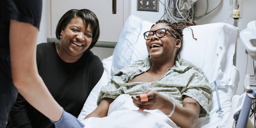 A young Black woman sitting in a hospital bed laughs with mother at her side. A nurse is in the foreground and medical equipment is on the wall behind the patient.