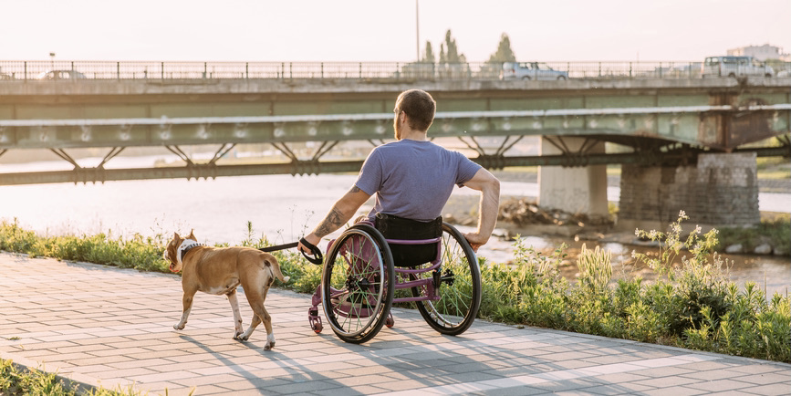 A young white man in a wheelchair rolls down a path along a river, with a dog on a leash walking beside him. 